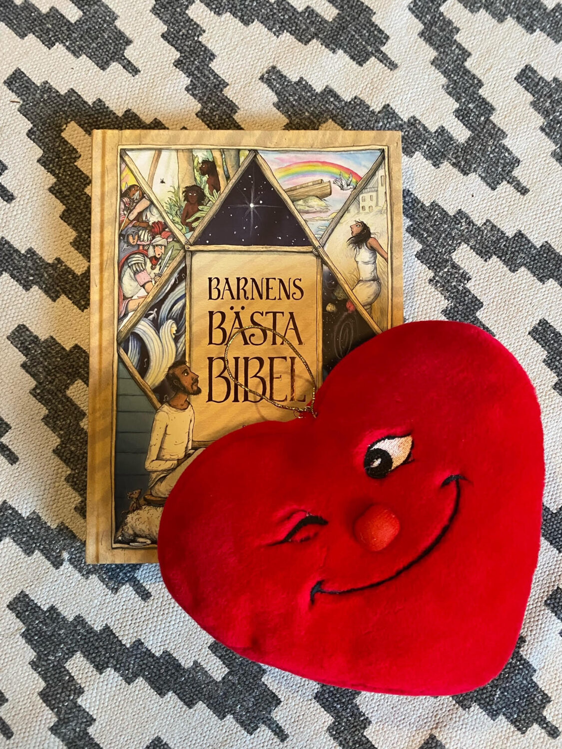 Barnkyrkans maskot Hjätrud: ett stort rött tyghjärta med mun, näsa och ögon, det ena ögat blinkar till oss. Bakom ligger Barnens bästa bibel.