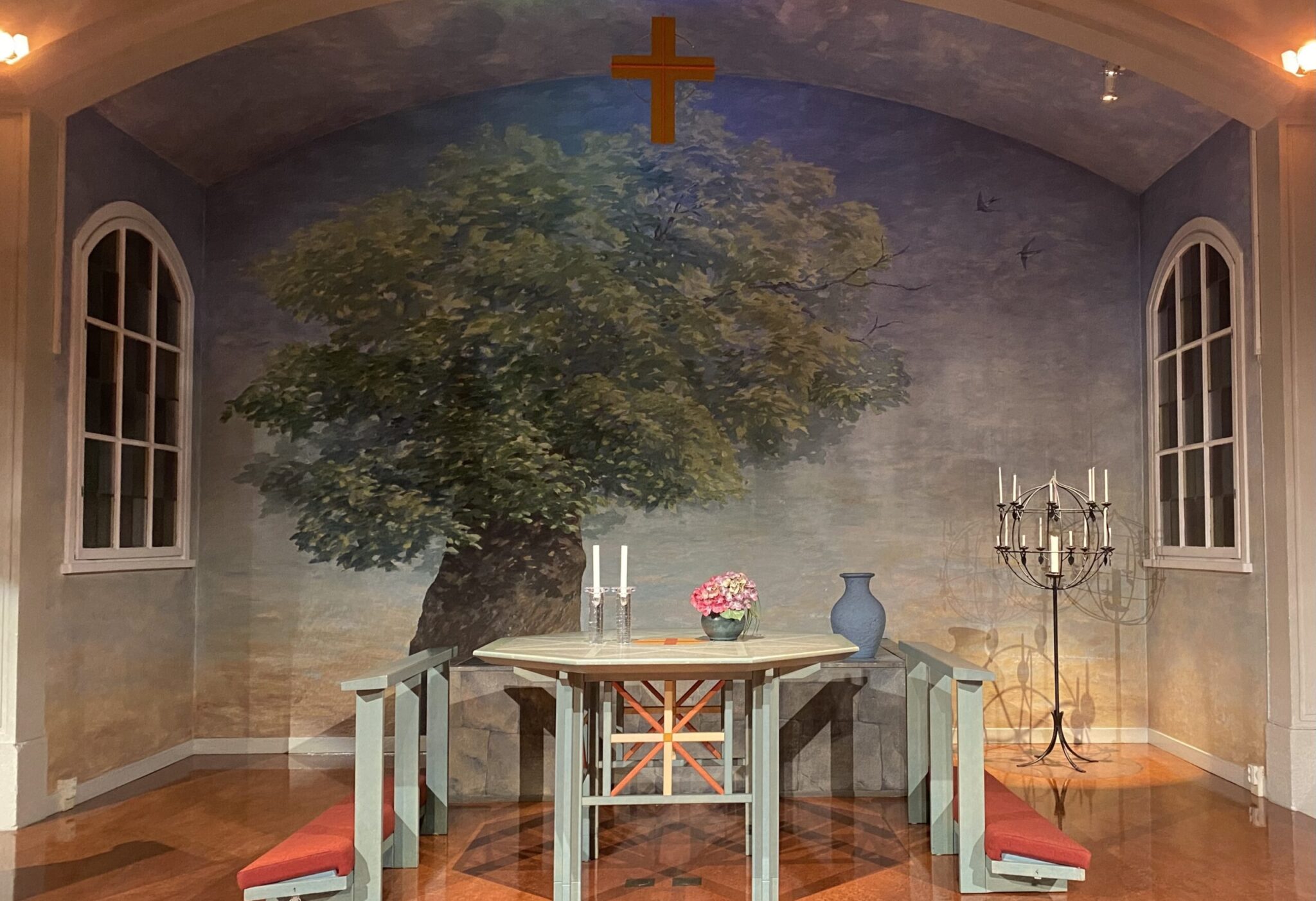 Altaret med ljus och blommor på. Till höger den stora ljusstaken och i bakgrunden altartavlan med ett stort träd med grön krona. I förgrunden i taket hänger ett rött kors.