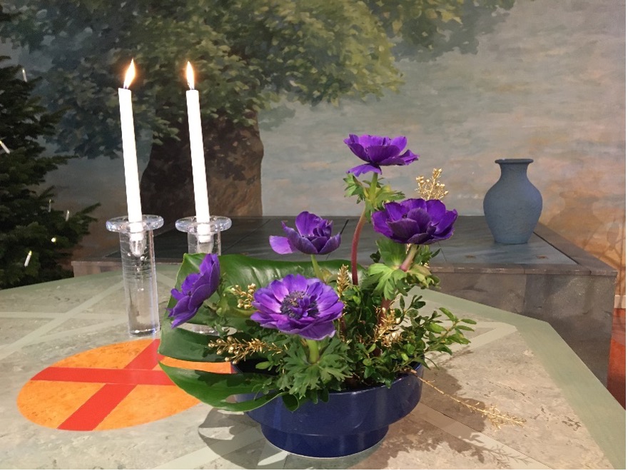 Altarbordet i Älvsjökyrkan, med två tända ljus och en växt med lila blommor, under syns en rundel i orange med ett mörkt orange kors på.