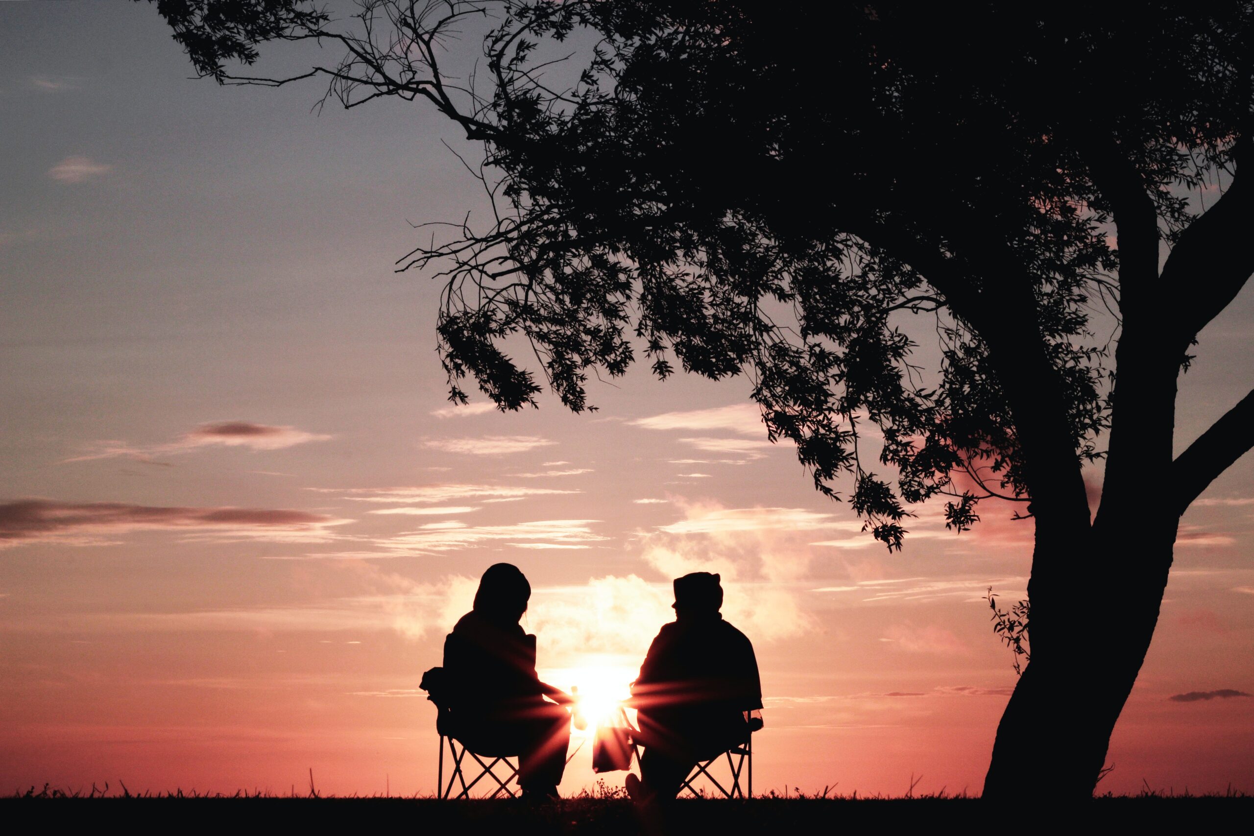 Två personer i silhuett sitter på varsin stol under ett träd och pratar med varandra. Solen går ner på en orange kvällshimmel och solen skiner mellan de två personerna.