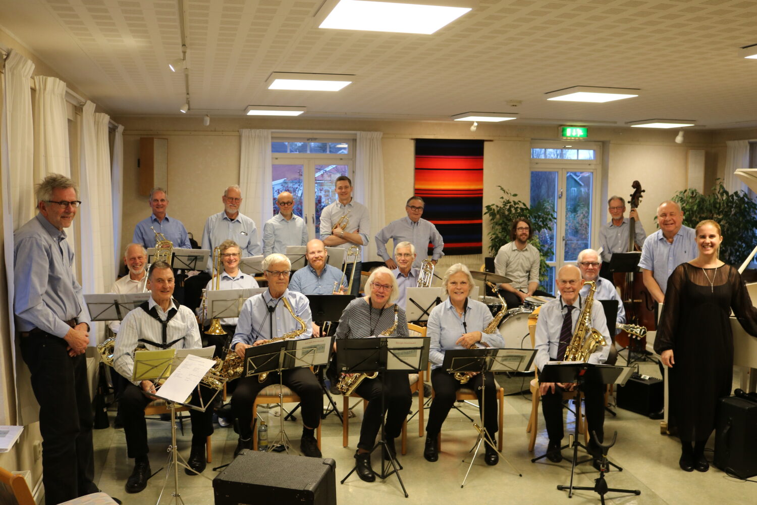 Hela Älvsjö storband sitter i sin replokal med instrument och notställ. Längst fram till vänster står deras dirigent och ler.
